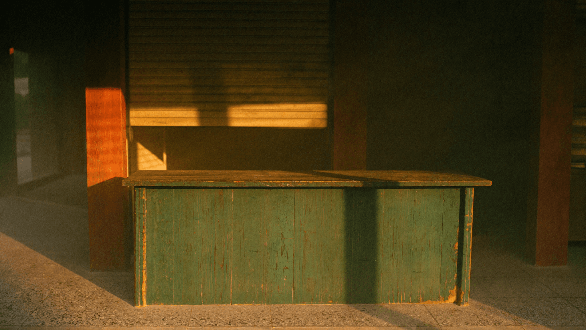 A shopfront counter of faded green painted timber on a terrazzo floor, lit by low golden morning light.